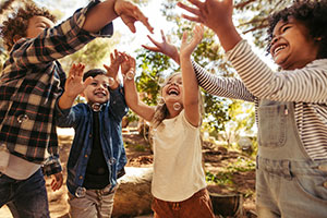 Vier Kinder stehen im Wald in einem Halbkreis und halten die Hände nach oben. Sie lachen, wirken sehr glücklich und spielen mit Seifenblasen.