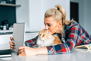 Eine blonde Frau sitzt an einem Tisch und hat ein Tablet vor sich aufgestellt. Ihren Kopf hat sie auf ihre Katze gelegt, die auch auf den Tabletbildschirm schaut. Neben der Frau liegt ein aufgeschlagenes Buch, im Hintergrund ist eine Küche zu erkennen.