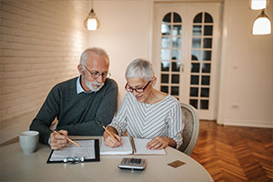 Ein älterer Mann und eine älteren Frau sitzen in ihrer Wohnung an einem Tisch und füllen Formulare aus. Auf dem Tisch befindet sich noch eine Tasse und ein Taschenrechner.