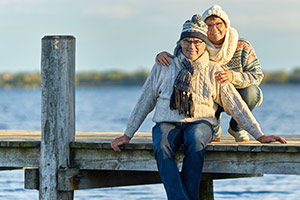 Ein Senioren-Pärchen sitzt auf einem Steg. Es ist Winter, die Kleidung ist warm. Im Hintergrund sieht man einen See, die Wintersonne scheint.