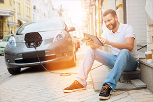 Ein junger Mann hält sitzend ein Tablet in der Hand während sein Elektrofahrzeug auflädt.