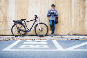 Ein älterer Mann steht auf einem Bürgersteig und lehnt sich mit dem Rücken an eine Wand, ein Bein ist angewinkelt. Er tippt etwas auf seinem Smartphone. Er trägt einen Fahrradhelm und eine Umhängetasche. Links neben ihm steht ein Elektro-Fahrrad. Auf der Straße vor ihm ist eine Spur mit der Aufschrift „BUS“ eingezeichnet.