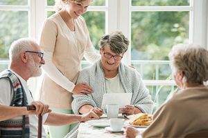 Eine Gruppe von drei Senioren sitzt an einem Frühstückstisch. Eine Seniorin ist von vorne zu sehen, sie hält ein Blatt Papier in der Hand und blickt lächelnd darauf. Eine junge Pflegerin steht neben ihr und legt ihr de Hand auf die Schulter.