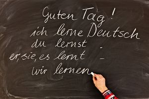 Eine Hand schreibt mit Kreide an eine Tafel „Guten Tag! Ich lerne Deutsch, du lernst, er, sie, es lernt, wir lernen“