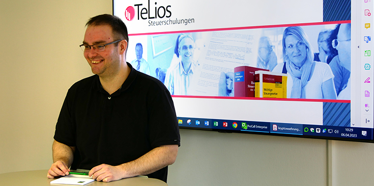 Der neue TeLios-Dozent Dominik Ferdinand steht vor einem Fernseher, auf dem Schulungsinhalte präsentiert werden.