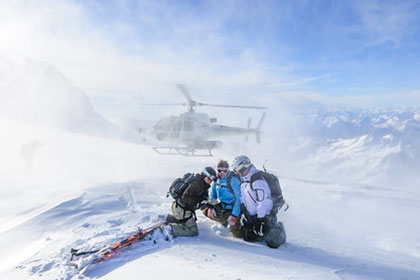 Oben angekommen, um die Abfahrt zu starten: Hartmut Gundlach und Freunde beim Heliskiing. Drei Personen in Ski-Montur sitzen auf einem schneebedeckten Berg und schauen auf eine Karte. Im Intergrund sind weitere schneebedeckte Berge, blauer Himmel und ein Helikopter zu sehen, der Schnee aufwirbelt.