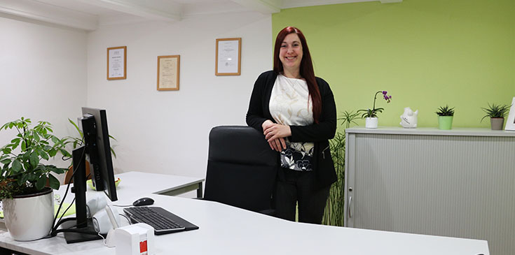 „Wer nicht wagt, der nicht gewinnt“ – Interview mit Berater-Neuling Carina Ebeling Eine Frau mit langen Haaren steht hinter ihrem Schreibtisch, an einem Schreibtischstuhl gelehnt und schaut lächelnd in die Kamera.