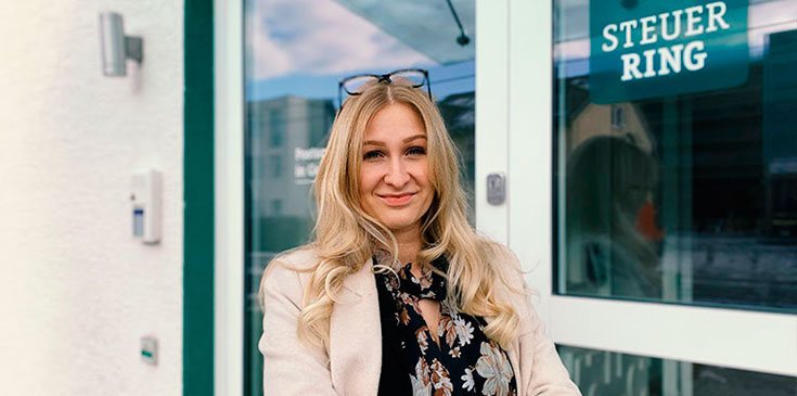 Eine blonde Frau in Bluse und Mantel steht vor einer Glastür. Sie trägt eine Brille auf dem Kopf und auf der Glastür ist das Steuerring-Logo erkennbar.