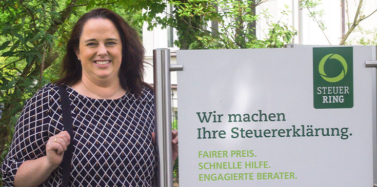 Kein Tag wie der andere: Interview mit Melanie Früchtenicht, Steuerring-Bereichsleiterin Eine Frau in gemusterter Bluse steht neben einem Schild, beschriftet mit Steuerring-Logo und „Wir machen Ihre Steuererklärung“.