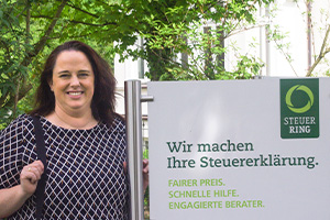 Eine Frau in gemusterter Bluse steht neben einem Schild, beschriftet mit Steuerring-Logo und „Wir machen Ihre Steuererklärung“.