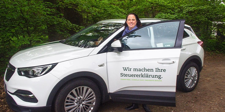 Treuer Begleiter: Mit ihrem Steuerring-Auto ist Melanie Früchtenicht in der Regel häufig unterwegs. Eine Frau steht in der Tür ihres Autos, das mit dem Steuerring-Claim „Wir machen Ihre Steuererklärung“ bedruckt ist.