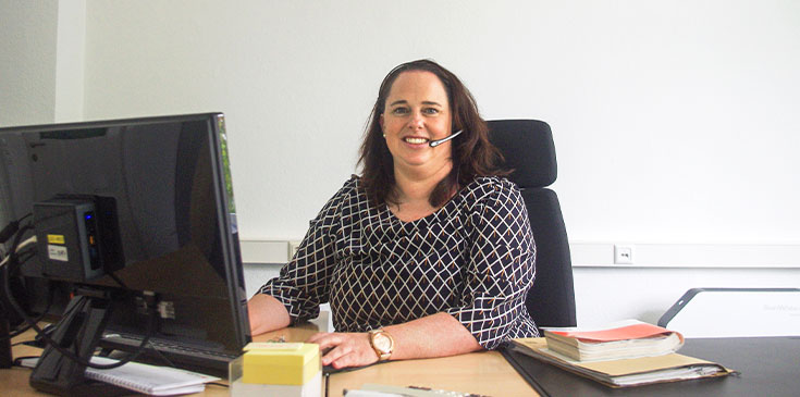 Melanie Früchtenicht analysiert genau, welche Art der Einarbeitung die Berater aus ihrem Bereich brauchen. Eine Frau mit gemusterter Bluse sitzt an ihrem Schreibtisch vor dem PC. Sie trägt ein Headset, auf dem Schreibtisch befinden sich Bücher und Unterlagen.
