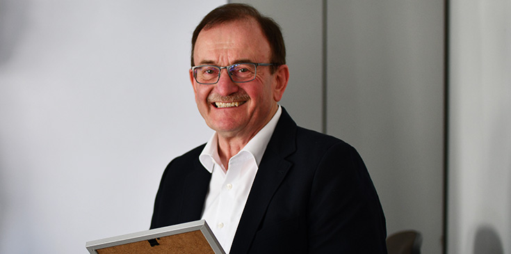 Ein Mann in Hemd und Jackett steht in einem Büro, lächelt und hält einen Fotorahmen in der Hand. Im Hintergrund befindet sich ein Schrank.