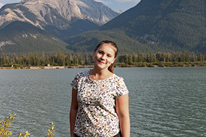 Eine junge Frau steht lächelnd vor einem See mit Bergen im Hintergrund.