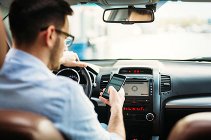 Fahrtkosten bei einer Auswärtstätigkeit richtig beantragen Ein Mann sitzt im Auto. Mit seiner linken Hand hält er das Steuer, in der rechten Hand hat er sein Handy. Man sieht den Rücken des Mannes und sein Profil.
