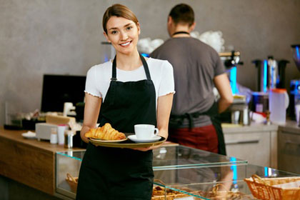 Ferienjobs: Wann für Schüler und Studenten Steuern fällig werden Eine junge Kellnerin mit blonden Haaren trägt eine schwarze Schürze und hält ein Tablett, auf dem ein Kaffee und ein Croissant platziert sind. Sie lächelt in die Kamera. Im Hintergrund steht ein Barista an der Kaffeemaschine.