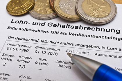 Prüfen Sie Ihre Lohnsteuerabzugsmerkmale Auf einer Lohn- und Gehaltsabrechnung sieht man die Lohnsteuerabzugsmerkmale, die geprüft werden sollten. Auf der Abrechnung liegen ein Kugelschreiber und Euro-Münzen.
