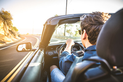 Auto, Arbeit, Urlaub?! Ein Mann in Shorts, hochgekrempeltem Hemd und mit Sonnenbrille fährt ein Cabrio um eine Straßenkurve. Der Mann ist von hinten zu sehen und die Sonne scheint auf ihn.