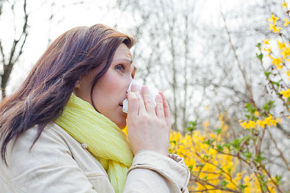 Allergien, Heuschnupfen: Medikamente und Therapien steuerlich absetzen Eine Frau mit rötlichen Haaren putzt sich mit einem leidvollen Gesicht die Nase. Im Hintergrund sieht man Bäume und Sträucher blühen.