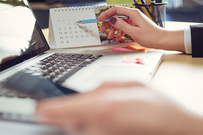 Stichtag 30.11.: Freibeträge beantragen und Steuererstattung für das ganze Jahr kassieren Von der Seite ist eine Hand zu erkennen, die einen Stift hält und auf ein bestimmtes Datum in einem Kalender zeigt. Dabei handelt es sich um einen Tischkalender, der auf einem Schreibtisch steht. Daneben ist ein Laptop platziert.