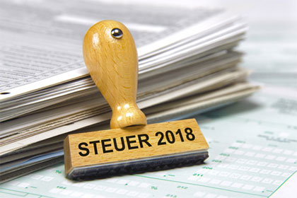Die Einkommensteuererklärung 2018 Ein Holzstempel mit der Aufschrift „Steuer 2018“ lehnt an einem Aktenstapel.