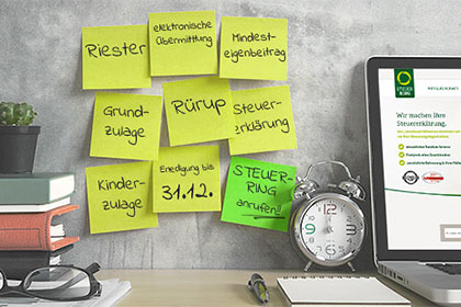 Riester-Rente, Rürup-Rente: Zulagen und Steuervorteile zum Jahresende sichern An einer Wand kleben neun Post-its, die unter anderem mit den Begriffen Riester, Rürup und Steuererklärung beschriftet sind. Rechts daneben befinden sich auf einem Tisch ein Wecker, ein Laptop und ein Block mit Stift. Links daneben sind Bücher gestapelt und es liegt eine Brille auf einem Stapel Papier.