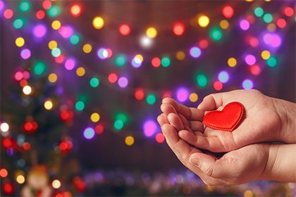 Spenden in der Weihnachtszeit, Bonus in der Steuererklärung Rechts im Bild liegen zwei geöffnete Hände übereinander und halten ein kleines Herz. Im Hintergrund sind verschwommen bunte Lichterketten platziert.