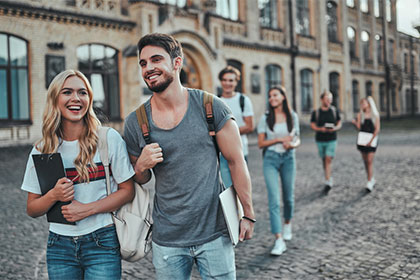 Steuererklärung während der Ausbildung Eine junge Frau und ein junger Mann laufen einer Gruppe voran auf einem Hof vor einem großen Gebäude entlang. Sie tragen Rucksäcke auf den Schultern und Unterlagen in den Händen. Beide lächeln und sehen zufrieden aus. Die restlichen Personen im Hintergrund sind verschwommen.