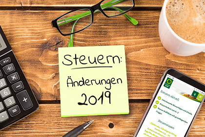 Ein gelber Notizzettel klebt auf einem Holztisch, darauf steht „Steuern: Änderungen 2019“. Um den Notizzettel herum liegen ein Taschenrechner, ein Kugelschreiber, eine Lesebrille und ein Smartphone mit der geöffneten Webseite des Steuerrings. Eine Tasse Milchkaffee rundet das Bild ab.