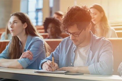 Sind Studienkosten trotz Stipendium von der Steuer absetzbar? In einem Vorlesungsaal sitzen Studenten, dabei sind zwei deutlich zu erkennen. Eine Studentin schaut nach vorne, ein anderer Student schreibt etwas auf. Der Hintergrund ist verschwommen.