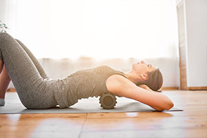 Eine junge Frau liegt auf einer Sportmatte auf dem Boden und hat die Beine angewinkelt. Sie rollt ihren Rücken über eine Rückenrolle und stützt mit ihren Händen den Kopf ab.