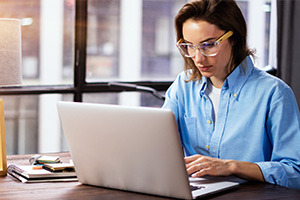 Eine junge Frau sitzt an einem Schreibtisch und schaut auf ihren aufgeklappten Laptop. Sie trägt eine Bluse und eine Brille. Auf dem Schreibtisch befinden sich Unterlagen und eine Tischlampe. Durch das große Fenster links im Bild scheint die Sonne und draußen sind verschwommen Häuser zu erkennen.