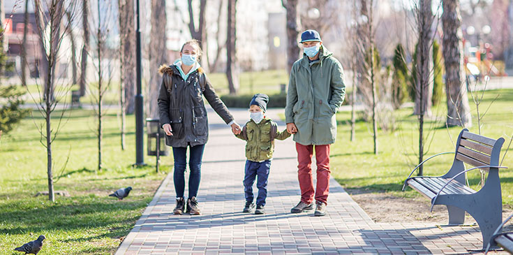Corona und die Folgen für die Steuererklärung Familienspaziergang zu Coronazeiten: Eine junge Frau und ein junger Mann halten in ihrer Mitte einen kleinen Jungen an den Händen und laufen auf einem gepflasterten Weg durch einen Park. Alle drei tragen eine Maske und blicken in die Kamera.