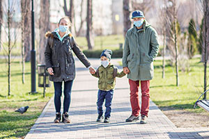 Familienspaziergang zu Coronazeiten: Eine junge Frau und ein junger Mann halten in ihrer Mitte einen kleinen Jungen an den Händen und laufen auf einem gepflasterten Weg durch einen Park. Alle drei tragen eine Maske und blicken in die Kamera.