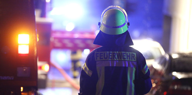 Ein Mann mit Helm und Feuerwehruniform ist von hinten zu sehen. Links neben ihm und verschwommen im Hintergrund sind Feuerwehrautos zu erkennen.