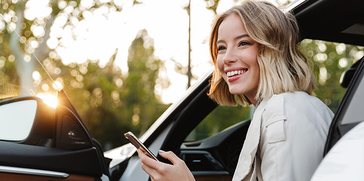 Eine Frau mit kurzen blonden Haare sitzt auf dem Fahrersitz ihres Autos. Sie hat die Tür geöffnet und schaut aus dem Auto heraus. Sie lächelt und hat ein Handy in der Hand.