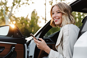 Eine Frau mit kurzen blonden Haare sitzt auf dem Fahrersitz ihres Autos. Sie hat die Tür geöffnet und schaut aus dem Auto heraus. Sie lächelt und hat ein Handy in der Hand.