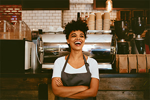 Eine Frau in T-Shirt und Schürze steht vor einer Kaffeetheke. Sie lacht, hat die Arme verschränkt und scheint eine Angestelltes des Cafés zu sein.