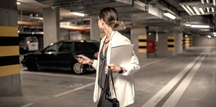 Homeoffice und Kurzarbeit: So wird der Firmenwagen günstiger Eine Frau in Blazer befindet sich in einer Tiefgarage. Sie dreht sich zu ihrem Auto um und schließt dieses mit einem Funkschlüssel zu. Sie hat in der anderen Hand eine Tasche und Unterlagen auf dem Arm.