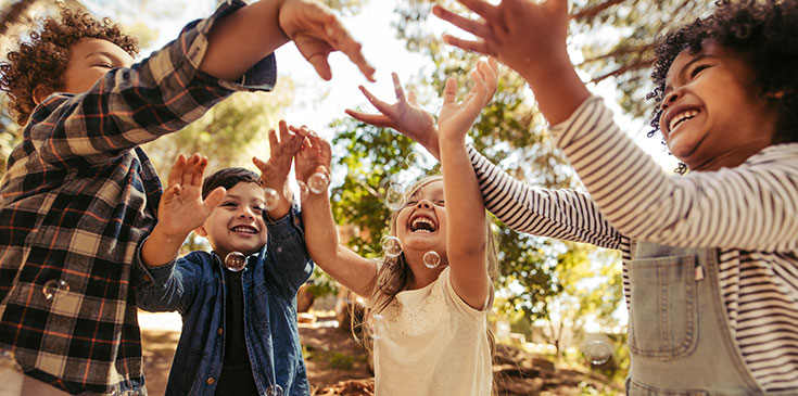 Kinderbetreuungskosten in der Steuererklärung absetzen Vier Kinder stehen im Wald in einem Halbkreis und halten die Hände nach oben. Sie lachen, wirken sehr glücklich und spielen mit Seifenblasen.