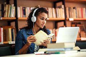 Eine junge Frau mit braunen, lockigen Haaren sitzt in der Bibliothek an einem Schreibtisch. Sie schaut auf den aufgeklappten Laptop und trägt Kopfhörer. In ihrer Hand hält sie ein Buch, auch um sie herum sind Bücher zu erkennen.