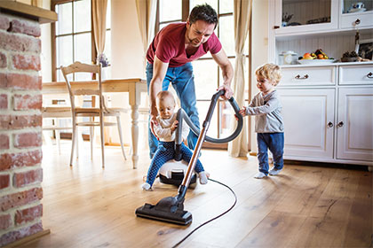 Minijob im Privathaushalt: Wie meldet man eine Haushaltshilfe an? Ein Vater in Jeans und T-Shirt befindet sich im Essbereich eines Hauses und bedient einen Staubsauer. Auf dem Staubsauger sitzt ein Baby und ein Kleinkind hält den Schlauch des Staubsauers fest. Der Mann hält das Baby fest; er lächelt zwar, wirkt jedoch leicht überfordert.