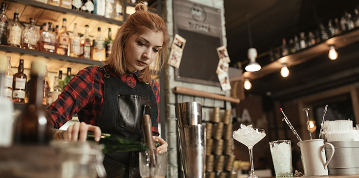 Ist ein Minijob steuerpflichtig? Eine Frau im karierten Hemd steht hinter einer Baar und mixt Cocktails. Sie hat einen kleinen Haardutt und trägt eine Schürze. Im Hintergrund sind verschwommen Getränkeflaschen, Gläser und Lichte zu erkennen.