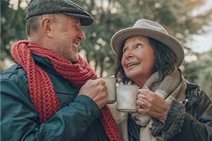 Auf dem Bild ist links ein älterer Mann und rechts eine ältere Dame zu erkennen. Sie tragen beide einen Schal und einen Hut. Sie schauen sich lächelnd in die Augen. Man kann erkennen, dass beide eine Tasse in der Hand halten und miteinander anstoßen.