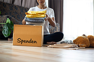 Eine Frau in T-Shirt und Jeans sitzt im Schneidersitz in ihrem Wohnzimmer und legt gefaltete Kleidung in eine Box mit der Aufschrift „Spenden“. Neben ihr liegen Kleiderbügel und ein Teddybär. Im Hintergrund sind eine Couch und eine Pflanze platziert.