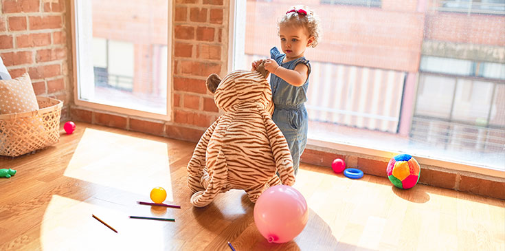 Ein kleines Kind steht im Kindergarten vor einem großen Fenster. Sie hält einen großen Stoffbären am Ohr. Um sie herum liegen Stifte, Luftballons und weitere Spielsachen.