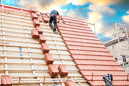 Steuervorteile für energiesparende Sanierungsmaßnahmen Ein Dachdecker arbeitet auf einem Dach und bringt die Dachziegel an. Er steht fast ganz oben auf dem Dach und ist von hinten abgebildet. Rechts oben im Bild befindet sich ein wolkenbedeckter Himmel.