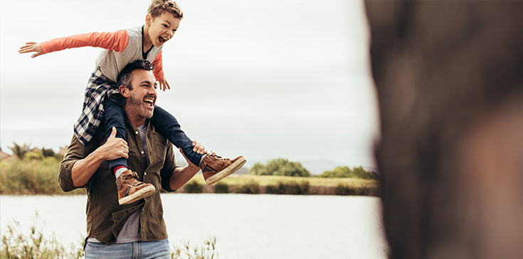 Ein Vater läuft an einem See entlang und trägt dabei seinen Sohn auf den Schultern. Beide lachen und freuen sich. Im Hintergrund sind verschwommen Sträucher zu erkenne.