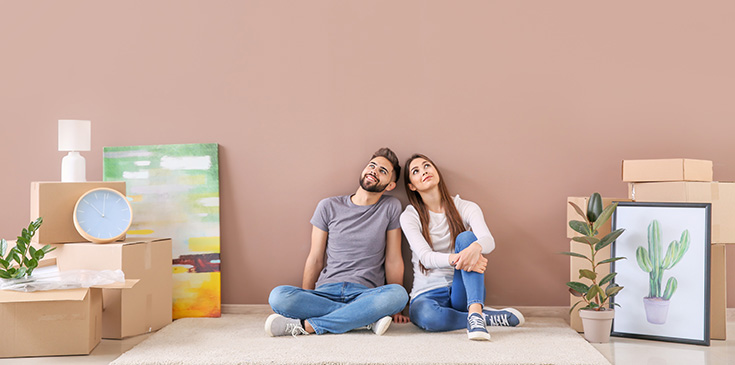 Umzugskosten in der Steuererklärung ansetzen Ein junger Mann und eine junge Frau sitzen auf dem Boden vor einer Wand in ihrer neuen Wohnung. Sie schauen nach oben, um sie herum stehen Umzugskisten, Bilder und Pflanzen.