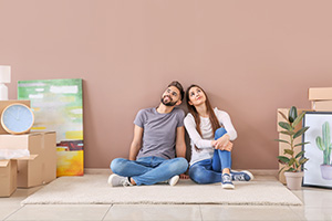 Ein junger Mann und eine junge Frau sitzen auf dem Boden vor einer Wand in ihrer neuen Wohnung. Sie schauen nach oben, um sie herum stehen Umzugskisten, Bilder und Pflanzen.